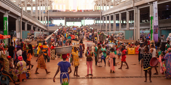Es ist ein Markt im globalen Süden zu erkennen. Es laufen Personen mit Gütern auf dem Kopf herum, welche sie balancieren