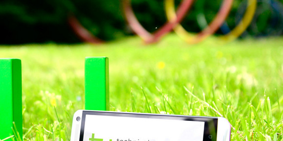 TU-App Ein grünes TU-Logo steht auf grünem Rasen. Daran lehnt ein Smartphone.